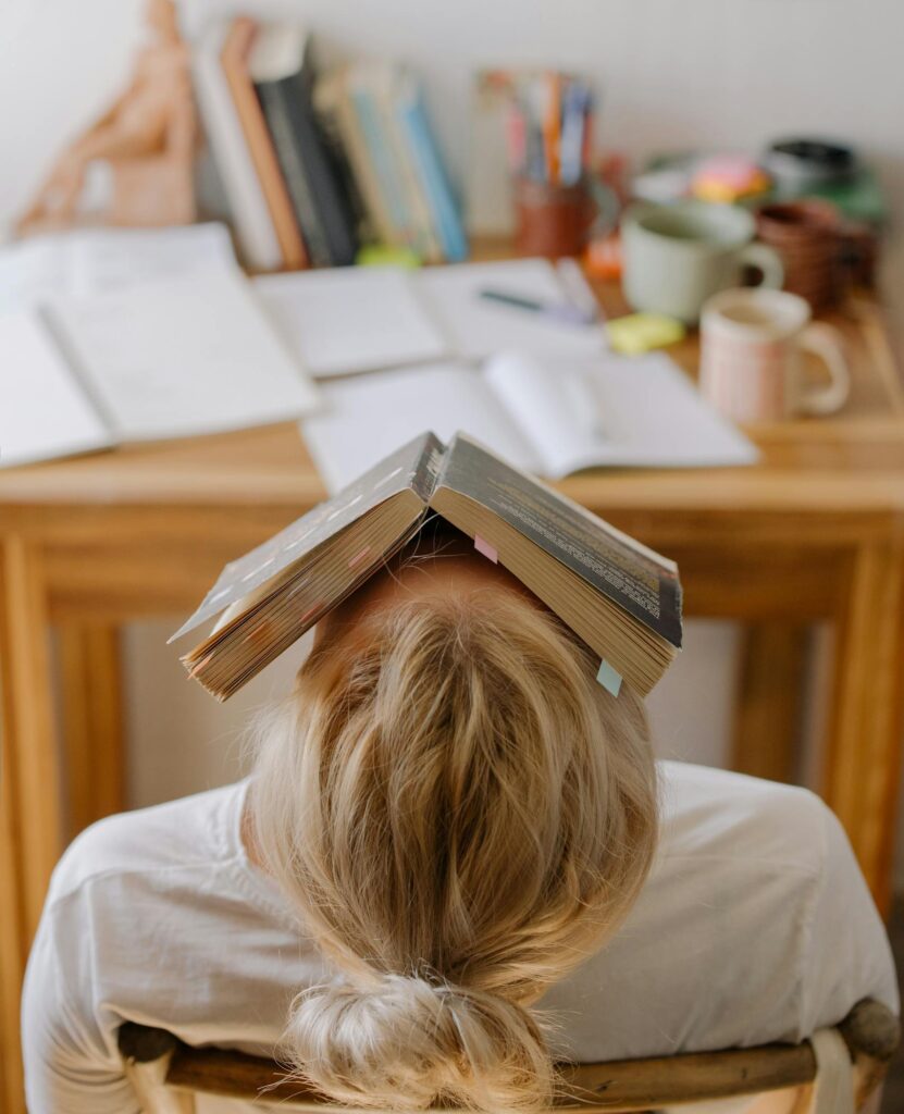 Student feeling stress and exhaustion while studying at a cluttered desk with an open book on their head.